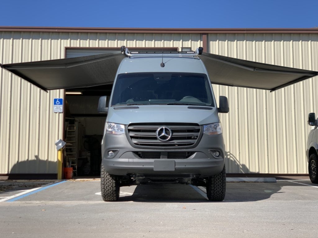 front of blue campervan with awning open on each side, parked in parking spot outside of yellow warehouse