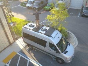 man standing on top of camper van with arms spread out - roof rack attached to top of van